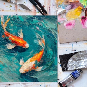 May include: Oil painting of two koi fish swimming in turquoise water. The fish are orange and white, with detailed brushstrokes. The painting is on a square canvas, with art supplies visible around it.