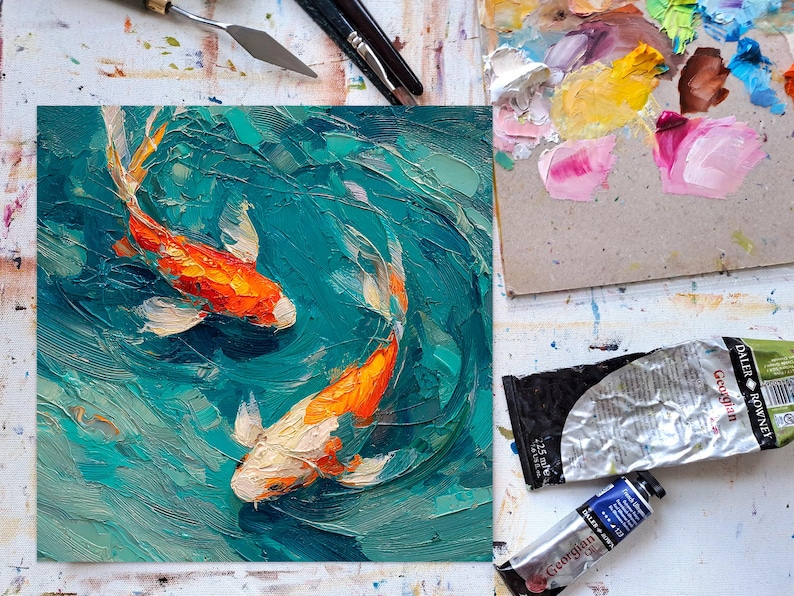 May include: Oil painting of two koi fish swimming in turquoise water. The fish are orange and white, with detailed brushstrokes. The painting is on a square canvas, with art supplies visible around it.