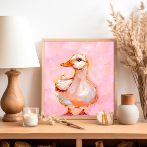 Op de afbeelding: Een schilderij van een witte eend met een roze achtergrond. De eend kijkt naar rechts en heeft een lichtoranje snavel. Het schilderij is ingelijst in een licht houten lijst.