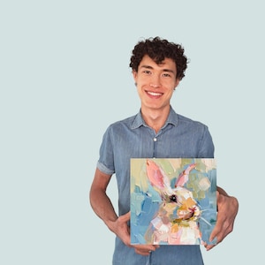 May include: A person holding a painting of a white rabbit with pink ears and a blue background. The rabbit is painted in a loose, impressionistic style.