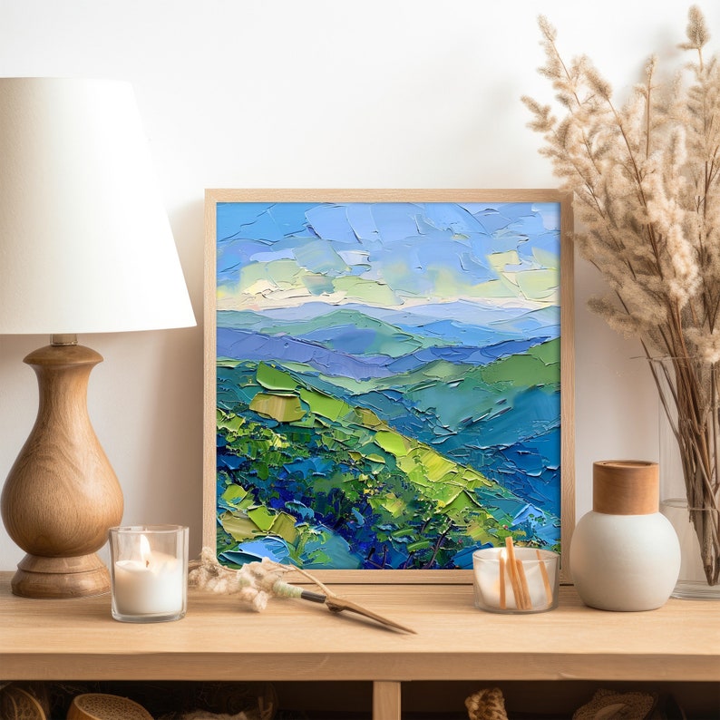 Op de afbeelding: Een ingelijst schilderij van een landschap met blauwe en groene bergen onder een blauwe lucht met witte wolken. Het schilderij is in een houten lijst en staat op een houten tafel.