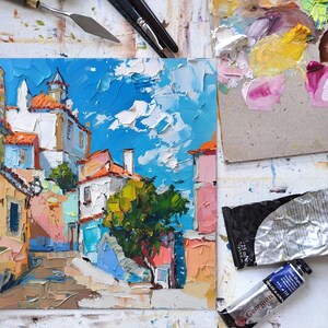 May include: A colorful oil painting of a narrow street in a European city. The buildings are painted in shades of yellow, orange, pink, and blue. There is a small tree in the center of the street. The sky is blue with white clouds.