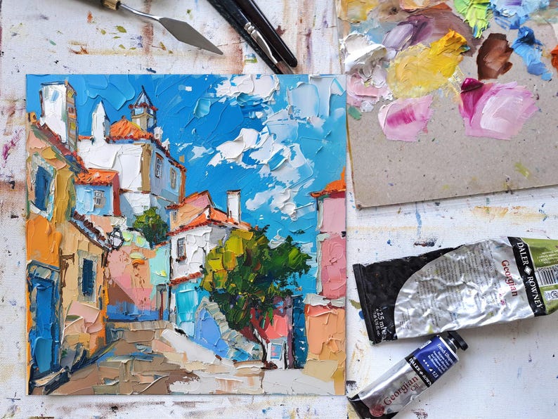 May include: A colorful oil painting of a narrow street in a European city. The buildings are painted in shades of yellow, orange, pink, and blue. There is a small tree in the center of the street. The sky is blue with white clouds.
