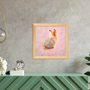 May include: A framed painting of a white rabbit with pink ears sitting on a pink background.