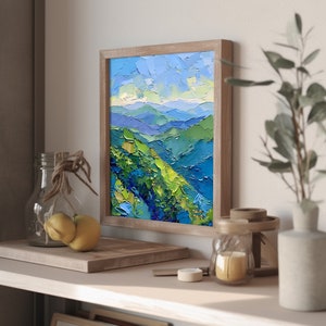 Op de afbeelding: Een ingelijst schilderij van een landschap met blauwe en groene bergen onder een blauwe lucht met witte wolken. Het schilderij is in een houten lijst.