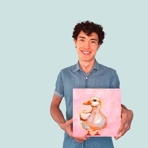 Op de afbeelding: Een persoon houdt een schilderij vast van een witte eend op een roze achtergrond. De eend kijkt naar rechts en heeft een gele snavel.