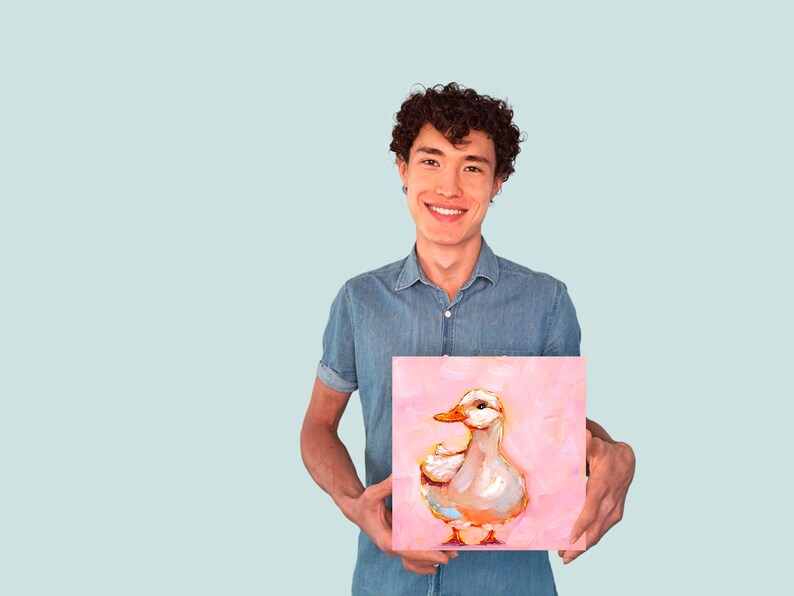 Op de afbeelding: Een persoon houdt een schilderij vast van een witte eend op een roze achtergrond. De eend kijkt naar rechts en heeft een gele snavel.