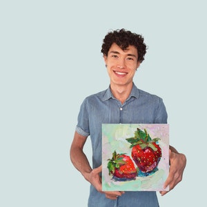 May include: A person holding a painting of two red strawberries with green leaves on a white canvas.