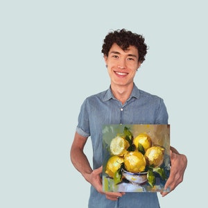 May include: A person holding a painting of a still life with lemons in a bowl. The lemons are yellow and the bowl is white with blue and green accents. The background of the painting is a light brown color.