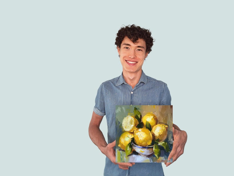 May include: A person holding a painting of a still life with lemons in a bowl. The lemons are yellow and the bowl is white with blue and green accents. The background of the painting is a light brown color.