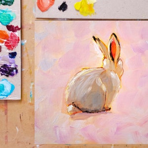 May include: A white rabbit with pink ears sits on a pink background. The rabbit is facing away from the viewer, with its tail visible. The background is painted with soft, brushstrokes.