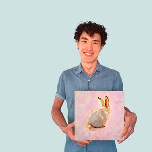 May include: A person holding a painting of a white rabbit with pink ears on a pink background.