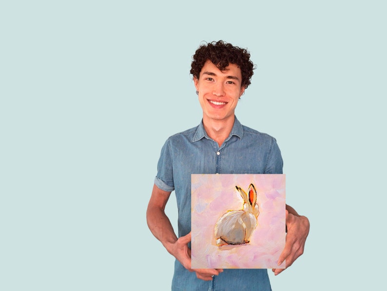 May include: A person holding a painting of a white rabbit with pink ears on a pink background.