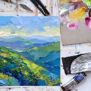 Op de afbeelding: Een close-up van een klein olieverfschilderij dat een landschap van glooiende groene heuvels en blauwe bergen onder een helderblauwe lucht afbeeldt. Het schilderij is gemaakt in een textuurrijke stijl, met zichtbare penseelstreken.