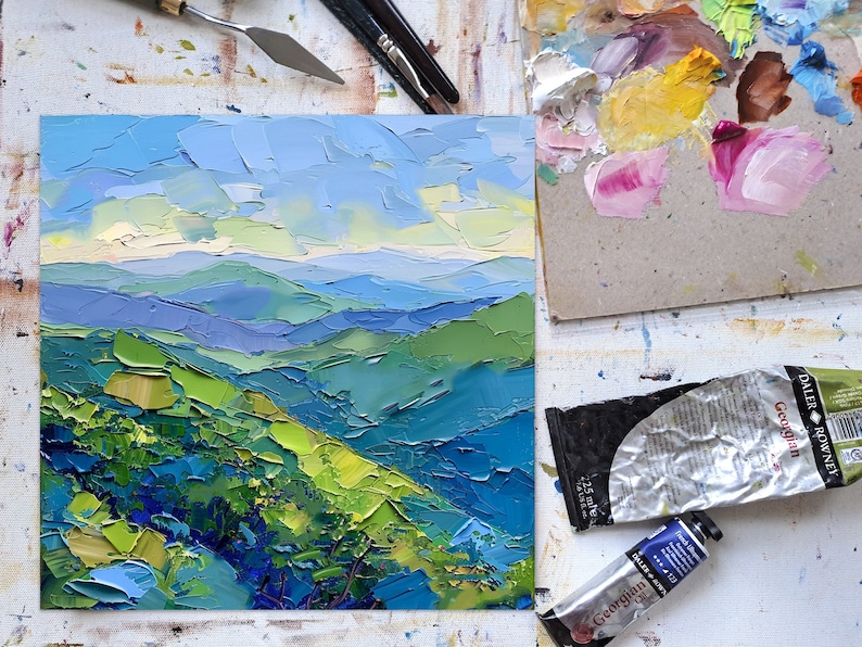 Op de afbeelding: Een close-up van een klein olieverfschilderij dat een landschap van glooiende groene heuvels en blauwe bergen onder een helderblauwe lucht afbeeldt. Het schilderij is gemaakt in een textuurrijke stijl, met zichtbare penseelstreken.