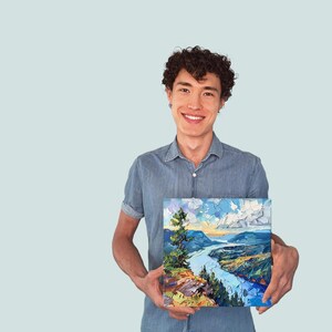 May include: A man in a blue shirt is holding a painting of a river winding through a valley with a mountain in the background. The painting has a blue sky with white clouds and a green tree in the foreground.