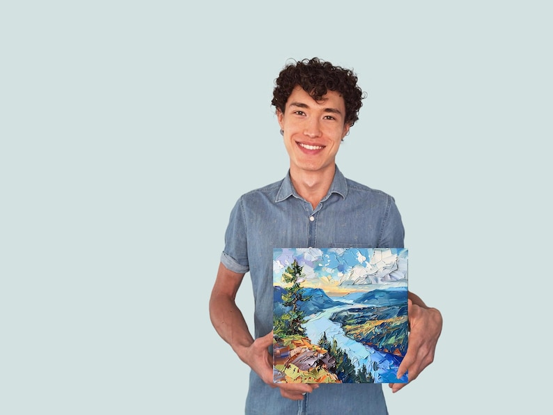 May include: A man in a blue shirt is holding a painting of a river winding through a valley with a mountain in the background. The painting has a blue sky with white clouds and a green tree in the foreground.