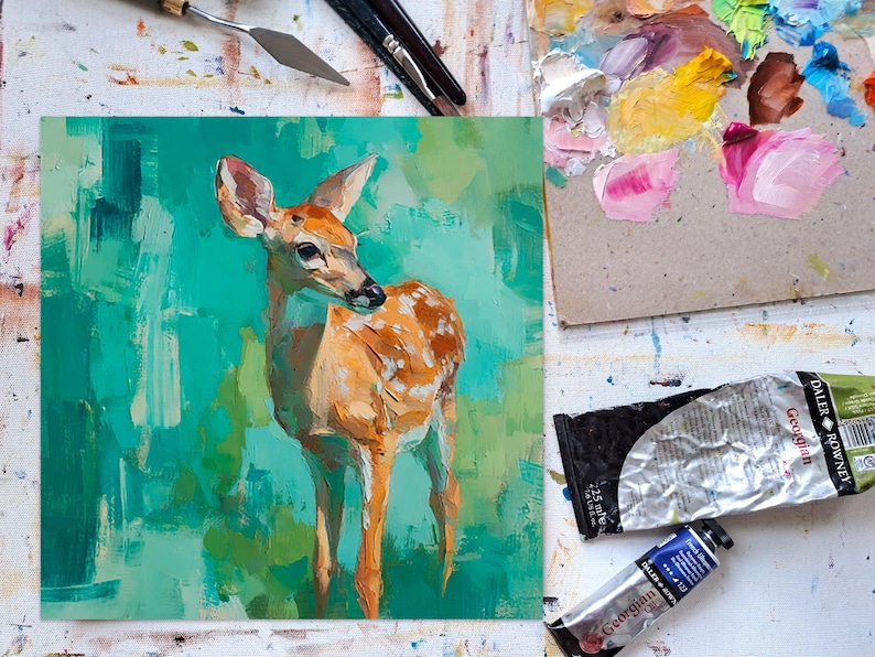 Puede incluir: Pintura al &oacute;leo original de un ciervo joven con pelaje marr&oacute;n claro y manchas blancas, sobre un fondo verde y turquesa. La obra de arte est&aacute; sobre una superficie blanca, con tubos de pintura, pinceles y una esp&aacute;tula visibles.