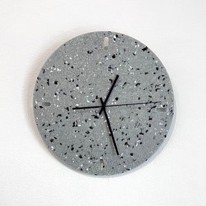 Puede incluir: Un reloj redondo gris con agujas negras y motas negras en la esfera. El reloj está colgado en una pared blanca. Un ramo de flores blancas está en un jarrón gris en la parte inferior de la imagen.