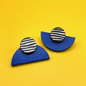 Puede incluir: Un par de pendientes geométricos. Cada pendiente presenta una forma de semicírculo en un vibrante color azul, coronado por un acento circular que muestra rayas horizontales en blanco y negro. Los pendientes están sobre un fondo amarillo brillante.
