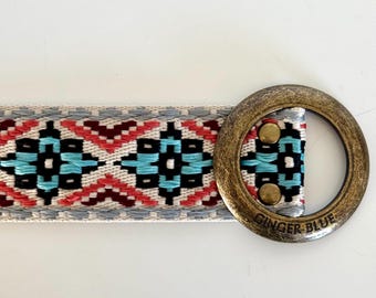 May include: A white woven belt with a geometric pattern in shades of red, blue, and black. The belt has a brass buckle with the words "Ginger Blue" engraved on it.