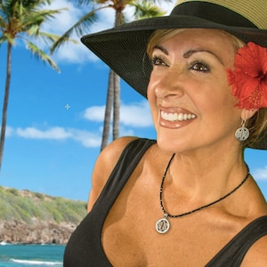 May include: A woman wearing a black sun hat with a red hibiscus flower, a black necklace with a silver pendant, and silver earrings. She is smiling and looking to the right. The background is a tropical beach scene with palm trees and blue sky.
