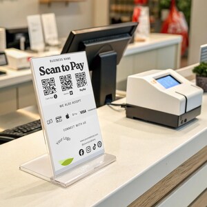 May include: A white and clear acrylic sign with the text "Scan to Pay" and QR codes. The sign also displays accepted payment methods and social media icons. A payment processing machine and a small potted plant are also visible.