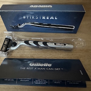 May include: A silver and black razor with a black handle and a silver blade. The razor is in a clear plastic package. The package is on a wooden surface. The razor has the word "Gillette" printed on it. The package has the text "#FIRSTREAL" printed on it.