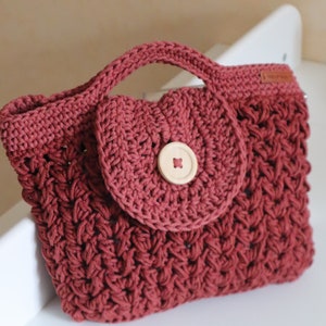 May include: A small, crocheted handbag in a deep red color with a round flap closure and a wooden button. The bag has a woven texture and a simple, classic design.
