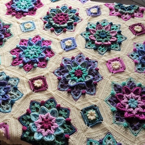 Könnte beinhalten: Eine gehäkelte Decke mit weißem Hintergrund und einem sich wiederholenden Muster aus bunten Blumen. Die Blumen sind in verschiedenen Blautönen, Lila, Rosa und Grün gearbeitet.