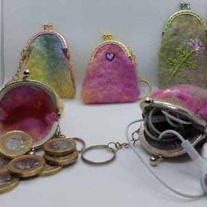 May include: A collection of handmade felt coin purses in various colors. The purses feature metal kiss-lock closures and keychains. One purse is open, displaying coins and another holds earbuds. The purses are in shades of pink, yellow, and green.