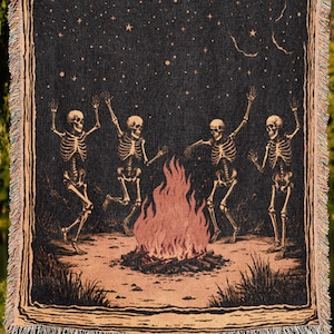 May include: A woven tapestry throw blanket with a black background and a design of four skeletons dancing around a campfire under a starry night sky. The text "Comfy Noodles woven throws and more" is printed below the blanket.