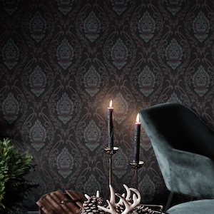 May include: A dark green velvet armchair with a black metal frame sits in a room with dark patterned wallpaper. A black metal side table with a wire basket holding two pine cones and a set of antlers sits in front of the chair. Two black candles are lit on a gold candlestick on the table.