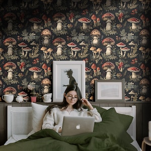May include: A woman sits on a bed with a green duvet cover, using a laptop computer. The wall behind her is covered in a dark wallpaper with a repeating pattern of red and white mushrooms and green foliage.