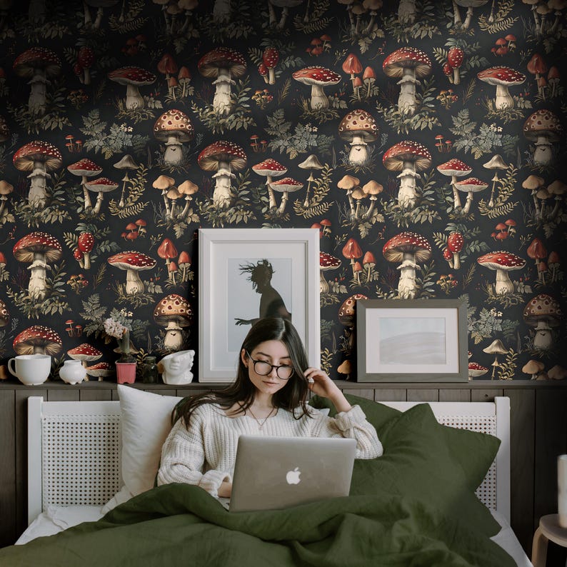 May include: A woman sits on a bed with a green duvet cover, using a laptop computer. The wall behind her is covered in a dark wallpaper with a repeating pattern of red and white mushrooms and green foliage.