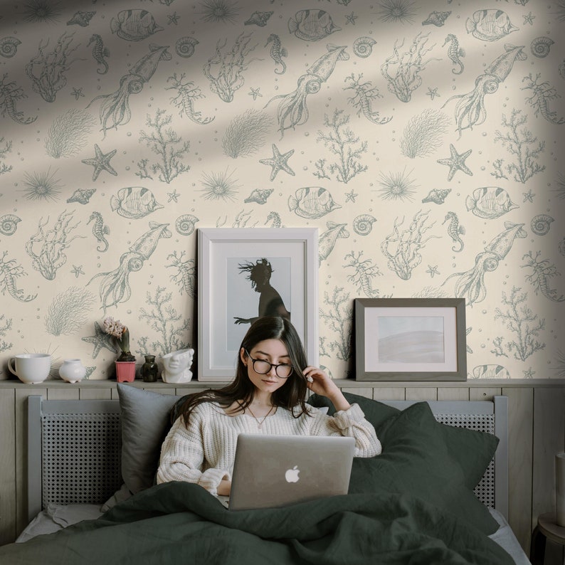 May include: A woman sits on a bed with a green headboard and a cream wallpaper with a repeating pattern of sea creatures, seashells, and coral. She is using a laptop computer with a white background and an apple logo.