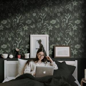 May include: A woman sits on a bed with a black and white floral patterned wallpaper, using a laptop computer. She is wearing a white sweater and glasses.