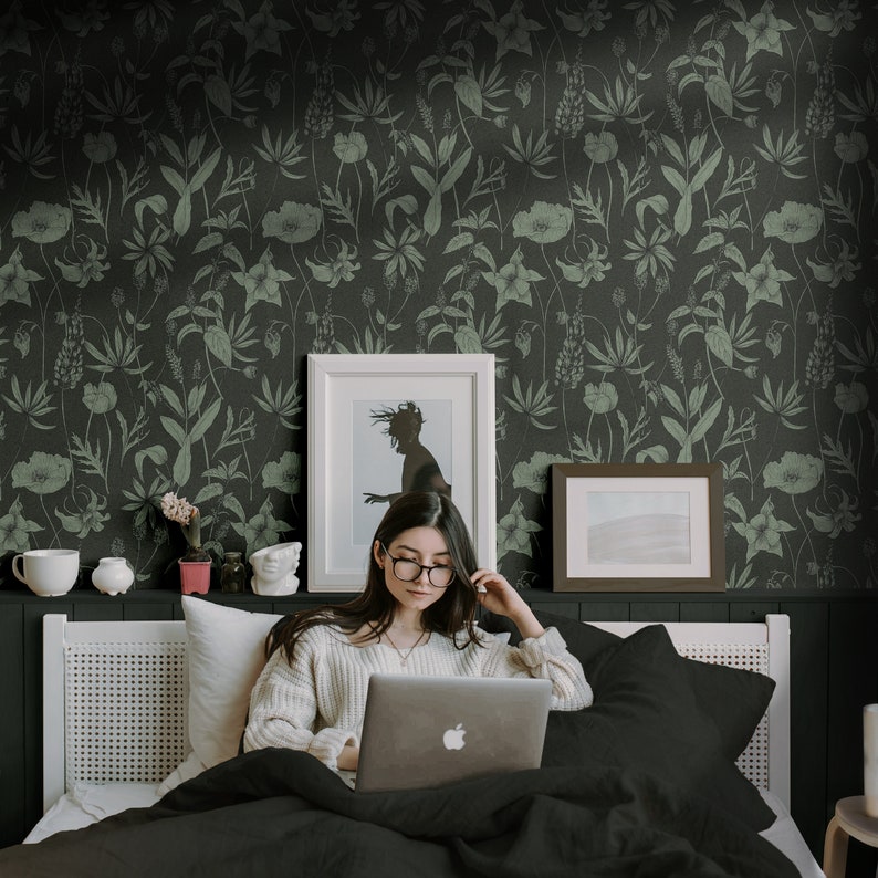 May include: A woman sits on a bed with a black and white floral patterned wallpaper, using a laptop computer. She is wearing a white sweater and glasses.