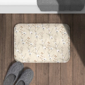 May include: A beige bath mat with a floral pattern of white flowers and brown stems. The mat is on a wooden floor.