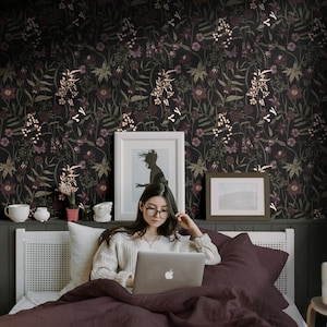 May include: A woman sits in bed with a laptop computer, a framed picture, and a framed photo on the wall behind her. The wall is covered in dark floral wallpaper with a black background and pink and white flowers.