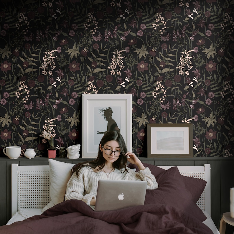 May include: A woman sits in bed with a laptop computer, a framed picture, and a framed photo on the wall behind her. The wall is covered in dark floral wallpaper with a black background and pink and white flowers.