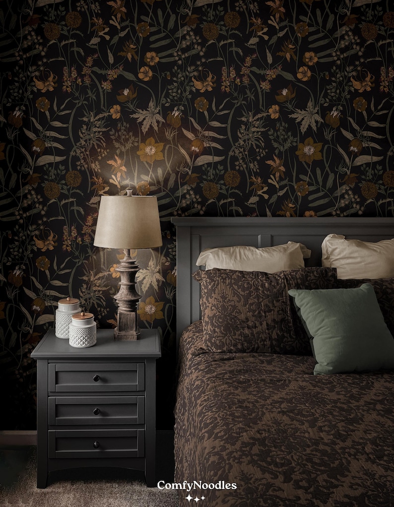May include: A dark gray nightstand with three drawers and black knobs. A lamp with a beige lampshade sits on the nightstand. The nightstand is next to a bed with a brown patterned duvet cover and pillows. The wall behind the bed is covered in dark floral wallpaper.