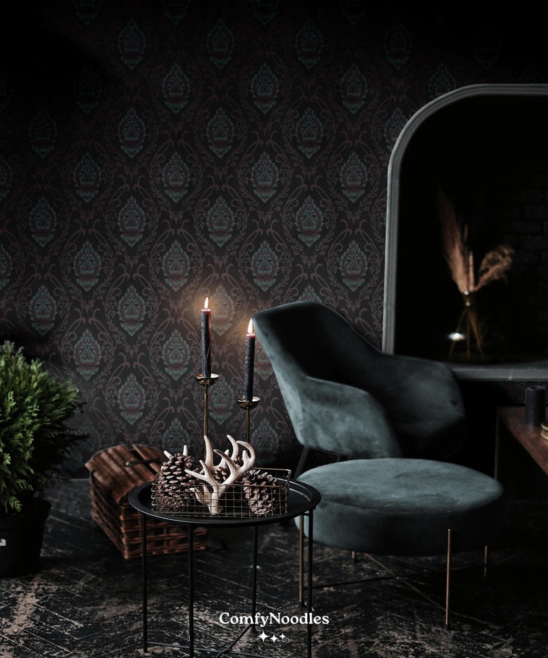 May include: A dark green velvet armchair with a black metal frame sits in a room with dark patterned wallpaper. A black metal side table with a wire basket holding two pine cones and a set of antlers sits in front of the chair. Two black candles are lit on a gold candlestick on the table.