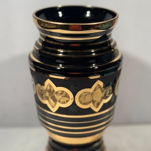 May include: A black vase with gold accents. The vase features horizontal gold bands and decorative gold floral patterns. The rim and base are also trimmed with gold. The vase has a glossy finish and a classic design.