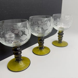 May include: Three clear wine glasses with etched grape and leaf designs. The stems are dark green with a spiral design, and the bases are a lighter green. The glasses are arranged in a row against a black and white background.