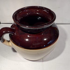 Könnte beinhalten: Ein zweifarbiges Steinzeuggefäß mit Henkel. Der obere Teil ist glänzend dunkelbraun, die untere Hälfte cremefarben beige. Das Gefäß hat eine weite Öffnung und einen abgerundeten Körper, geeignet für Küche oder Dekoration. Ein klassisches Stück.