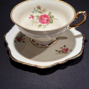 Puede incluir: Un juego de taza y platillo de té vintage. La taza y el platillo de porcelana blanca presentan un diseño floral de rosas rosas con hojas verdes. Los bordes y el asa están adornados con dorado. El platillo tiene un borde festoneado.