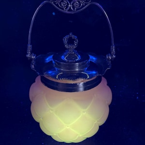 Puede incluir: Una mantequera vintage de cristal amarillo con asa y tapa plateadas. El plato tiene un diseño acolchado y un borde plateado. El asa es ornamentada con adornos decorativos. La mantequera está sobre una superficie oscura.