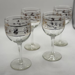 May include: Four clear wine glasses with a gold and black floral design around the rim. The glasses have a slender stem and a round base. The design includes pine needles and pine cones.