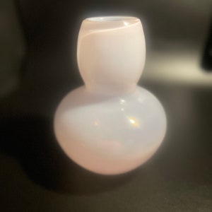 Peut inclure: Un petit vase en verre rose pâle, avec une base bulbeuse et un sommet arrondi plus étroit. Le vase a une finition lisse et brillante et un motif subtil en spirale visible dans le verre. La forme générale rappelle une gourde.